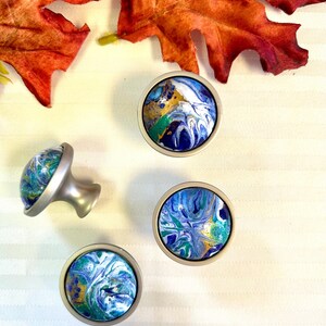 May include: Four decorative cabinet knobs with a marbled design in shades of blue, green, white, and gold. The knobs have a silver-colored base and are arranged on a white striped surface. Two orange autumn leaves are in the background.