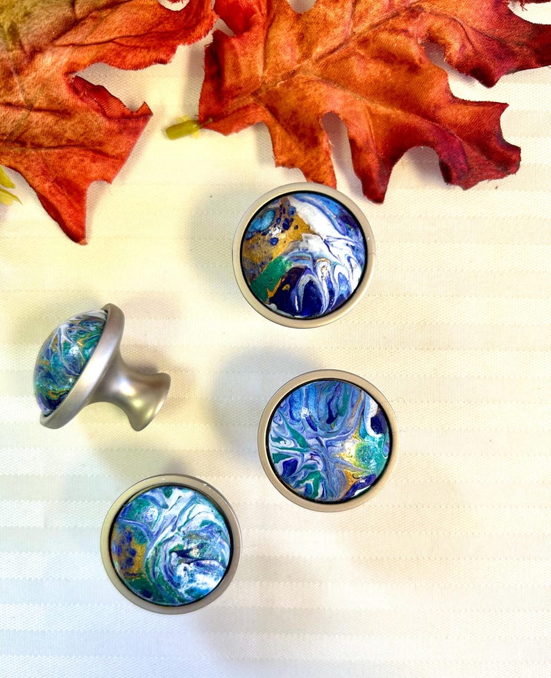 May include: Four decorative cabinet knobs with a marbled design in shades of blue, green, white, and gold. The knobs have a silver-colored base and are arranged on a white striped surface. Two orange autumn leaves are in the background.