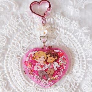 以下が含まれることがあります： 2 人の漫画のイラストが描かれた、ピンク色のハート型のキーホルダー。 キーホルダーにはハート型の留め金と、リボンとハートのチャームが付いた装飾チェーンが付いています。 「Happy Valentine's」の文字がハートに書かれています。