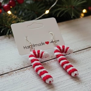 May include: A pair of handmade candy cane earrings. Each earring is a miniature candy cane design with red and white stripes. The earrings are displayed on a white card with the text "Handmade with love".