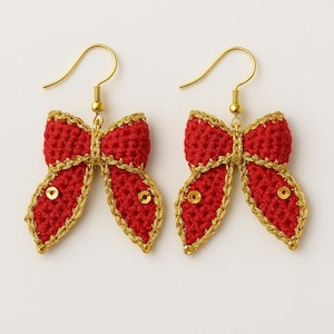 May include: A pair of red crochet bow earrings with gold trim and gold-coloured ear wires. Each bow features a small gold sequin in the centre. The earrings are displayed against a white background.
