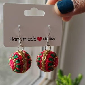 May include: A pair of handmade red and green embroidered ball earrings with gold accents. The earrings are suspended from silver-colored hooks and are displayed on a white card that reads "Handmade with love."