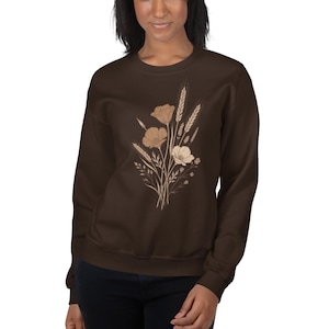 May include: A dark brown crewneck sweatshirt featuring a central graphic illustration of a botanical bouquet. The design consists of light beige line art depicting various wildflowers, grasses, and wheat stalks arranged in a vertical cluster.
