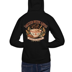 May include: A black hoodie featuring a back graphic of a bowl of porridge with a wooden spoon, wheat stalks, and cinnamon sticks. The text reads "MIXED WITH LOVE?" at the top and "DUSTED WITH CINNAMON" on a banner at the bottom in a warm colour palette.