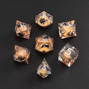 Op de afbeelding: Set van zeven heldere harsdobbelstenen met oranje en zwarte accenten. Elke dobbelsteen heeft een ander nummer en een zwarte kattenillustratie. De dobbelstenen zijn ontworpen voor tabletop gaming en zijn gerangschikt op een donkere ondergrond.