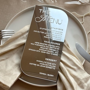 Könnte beinhalten: Eine reflektierende silberne Menükarte mit abgerundeten Kanten, die den Text "THE MENU" in eleganter Schrift zeigt. Die Speisekarte listet Vorspeisen, Hauptgerichte und Dessertoptionen auf. Die Karte liegt auf einem weißen Teller mit einer gefalteten beigen Serviette.