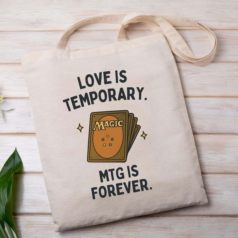 K&ouml;nnte beinhalten: Eine wei&szlig;e Canvas-Tragetasche mit schwarzem Text, der "Liebe ist verg&auml;nglich. MTG ist f&uuml;r immer." lautet. Die Tasche zeigt ein Cartoon-Bild einer Magic: The Gathering-Karte.