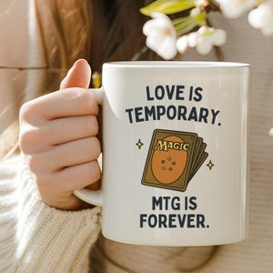K&ouml;nnte beinhalten: Wei&szlig;e Keramiktasse mit dem Text "Liebe ist verg&auml;nglich. MTG ist f&uuml;r immer." und einem Cartoon-Bild einer Magic: The Gathering Karte.