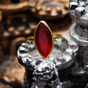 May include: A gold ring with a large, deep red, marquise-shaped gemstone. The ring is sitting on a small, gray, stone structure.