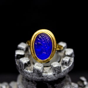 May include: A gold ring with a blue oval-shaped gemstone. The gemstone has a carved design and is set in a gold bezel.