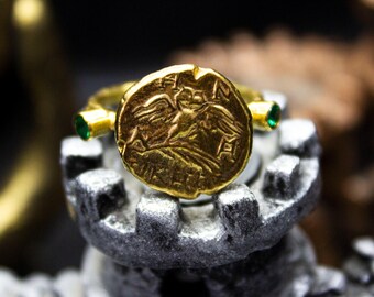 May include: A gold ring with a detailed coin design featuring a winged figure. The ring is accented with two emerald green gemstones.