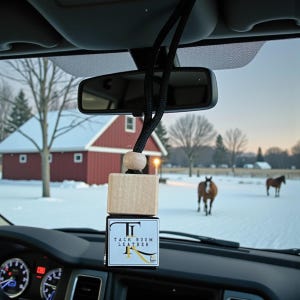 May include: An air freshener hanging from a car's rearview mirror. The air freshener is a wooden cube with the text "Tack Room Leather" on it. The background shows a snowy landscape with horses and a red barn.