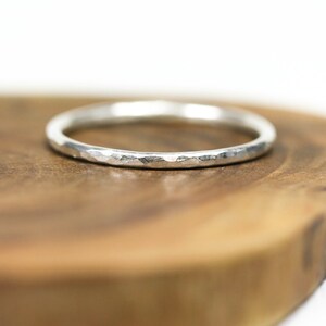 Può includere: Un anello in argento martellato con una superficie strutturata. L'anello è posto su uno sfondo di legno, evidenziando la sua lucentezza metallica. Il design dell'anello presenta una serie di piccole sfaccettature irregolari, creando un aspetto unico e accattivante.