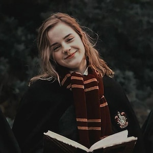May include: A person wearing a black robe and a burgundy and gold striped scarf, holding an open book. The scarf has a crest on it. The person is smiling, with a dark green background.