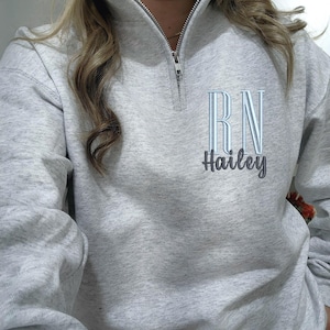 May include: A light grey heathered quarter-zip sweatshirt featuring an embroidered design on the left chest. The embroidery includes the letters RN in light blue and the name Hailey in dark grey script lettering below the letters.