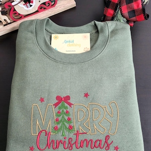 May include: An olive green sweatshirt with the embroidered words "MERRY Christmas" in gold and pink. A Christmas tree with red ornaments and a bow is in the center of the word "MERRY". The sweatshirt is folded neatly.