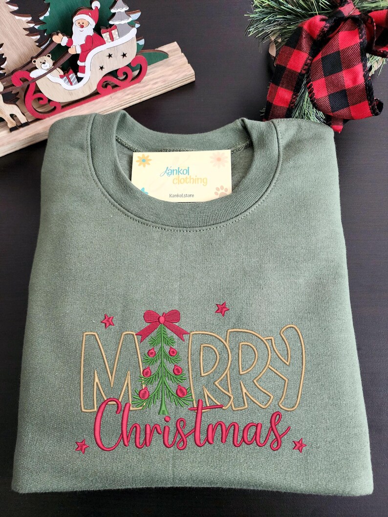 May include: An olive green sweatshirt with the embroidered words "MERRY Christmas" in gold and pink. A Christmas tree with red ornaments and a bow is in the center of the word "MERRY". The sweatshirt is folded neatly.