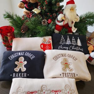 May include: A festive Christmas display featuring sweatshirts with gingerbread man designs and text. The sweatshirts are in navy and cream colors. A plush reindeer and Santa Claus figurine are also present, along with a decorated Christmas tree.