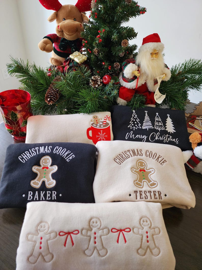 May include: A festive Christmas display featuring sweatshirts with gingerbread man designs and text. The sweatshirts are in navy and cream colors. A plush reindeer and Santa Claus figurine are also present, along with a decorated Christmas tree.