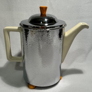 May include: A silver and white ceramic teapot with a hammered metal body and an orange plastic knob on the lid. The teapot has a white ceramic spout and handle.