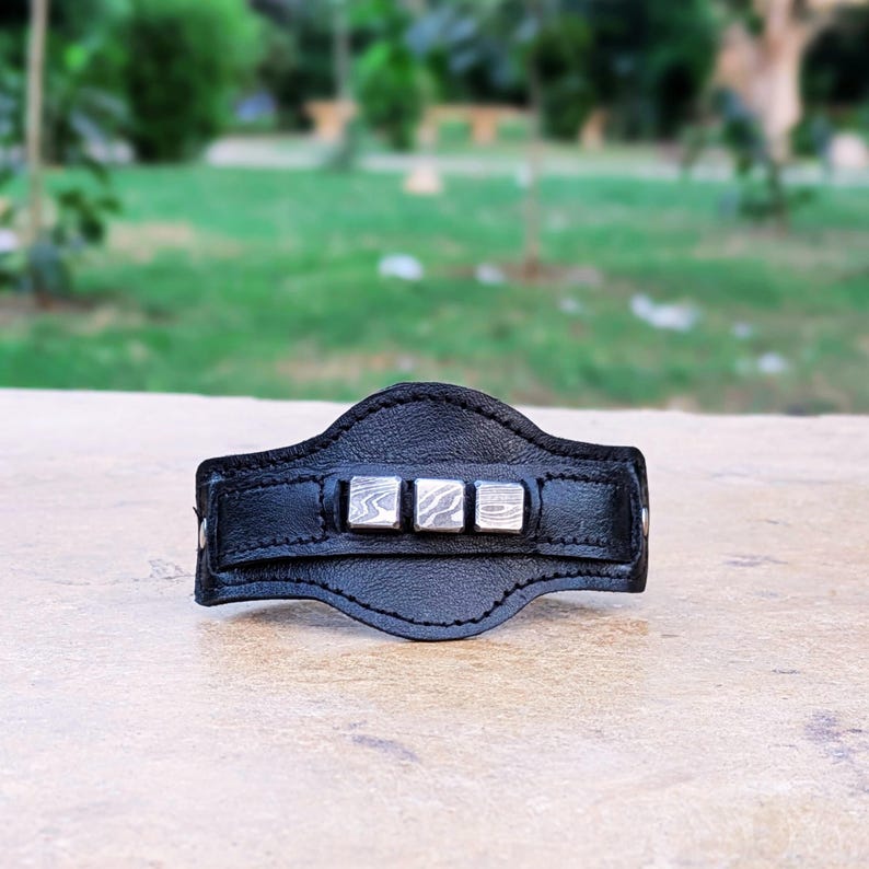 May include: Black leather bracelet with five silver square accents. The bracelet is on a wooden surface.
