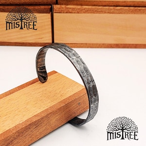 Könnte beinhalten: Ein gemustertes Metall-Armreif-Armband mit geschwungenem Design. Das Armband wird auf einem Holzblock präsentiert. Das Bild enthält das "Mistree"-Logo mit einem Baumdesign und Text.
