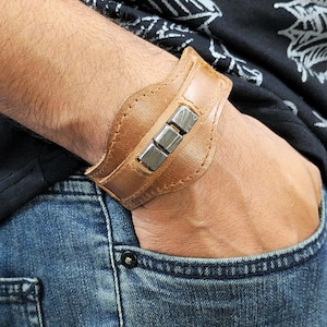 May include: A brown leather cuff bracelet with silver metal accents. The bracelet is worn on a person's wrist.