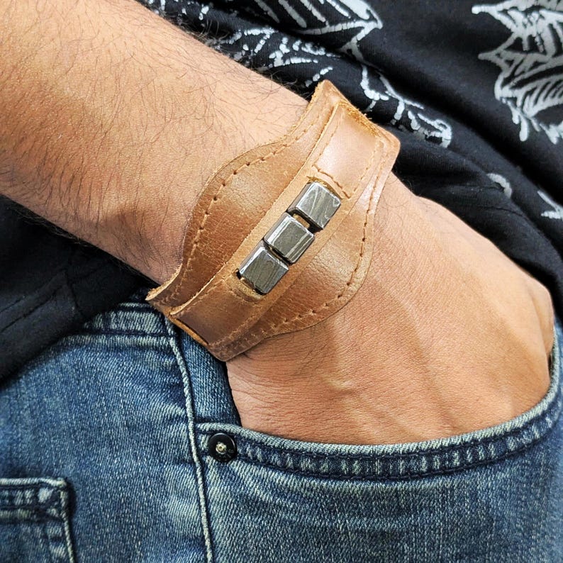 May include: A brown leather cuff bracelet with silver metal accents. The bracelet is worn on a person's wrist.