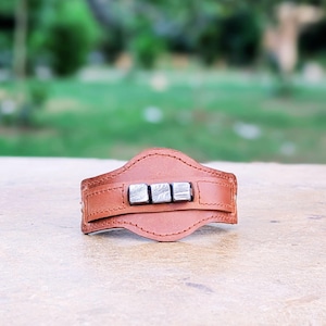 May include: A brown leather bracelet with four silver metal squares in the center.