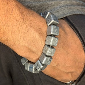 May include: A bracelet made of silver-toned metal with a geometric pattern. The bracelet is made of multiple square-shaped beads.