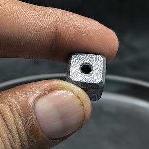 Könnte beinhalten: Eine kleine, quadratische Metallperle mit einem Loch in der Mitte. Die Perle hat ein wirbelndes, gemustertes Design.