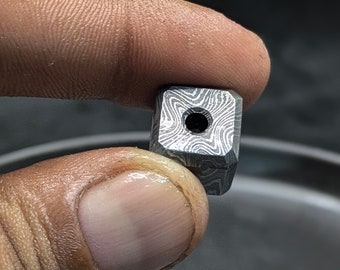 May include: A small, square, metal bead with a hole in the center. The bead has a swirling, patterned design.