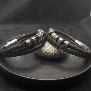 May include: Two leather bracelets, one black and one brown, with silver-coloured metal accents and magnetic clasps. The bracelets are braided and feature decorative beads with Celtic knot designs. They are displayed on a dark plate.