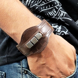 May include: A brown leather cuff bracelet with a silver metal accent. The metal accent is made of four rectangular pieces with a hammered finish.