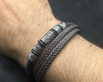 May include: A set of three bracelets. One is braided brown leather, another is black leather with silver-colored beads, and the third is black leather with silver-colored accents. The bracelets are stacked on a wrist.