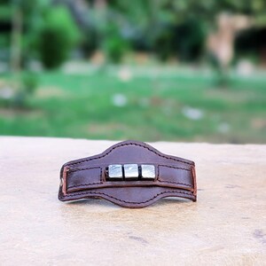 May include: A brown leather bracelet with a silver metal accent. The metal accent is made up of three square pieces with a textured surface.
