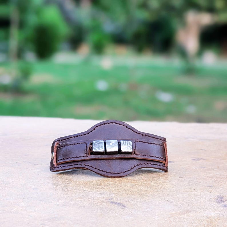 May include: A brown leather bracelet with a silver metal accent. The metal accent is made up of three square pieces with a textured surface.