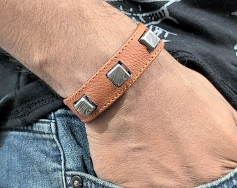May include: A brown leather bracelet with three silver square metal accents. The bracelet is worn on a person's wrist.