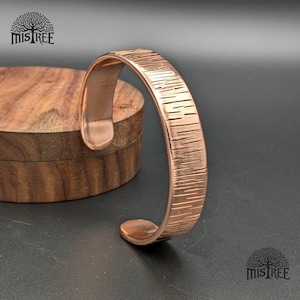May include: A copper cuff bracelet with a textured, linear pattern. The open-ended bracelet is displayed on a dark surface next to a wooden box. The bracelet's design features a series of parallel lines, creating a unique, tactile surface.
