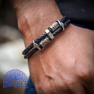 May include: Black leather bracelet with silver metal detailing. The bracelet has a braided leather design with decorative metal pieces. The bracelet is worn on a person's wrist.