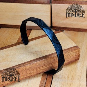 May include: A black metal cuff bracelet with a textured, patterned surface. The bracelet has a geometric design and is displayed on a wooden surface. The brand name "Mistree" is visible.