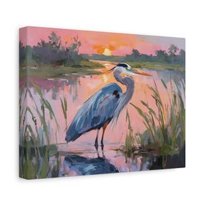 May include: Canvas art print of a great blue heron standing in water at sunset. The heron is depicted in shades of blue and gray, with an orange beak. The background features a pink and orange sky, water, and green reeds.