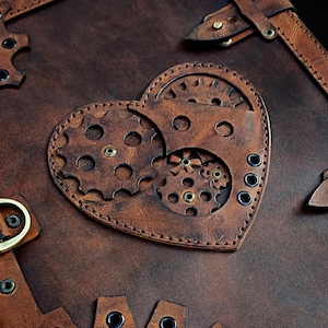 Può includere: Una copertina per diario in pelle marrone scuro con un ritaglio a forma di cuore sul davanti. Il cuore è riempito con ingranaggi in pelle marrone stratificati e rivetti metallici. La copertina presenta bordi cuciti e occhielli in metallo.