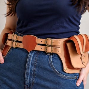 Op de afbeelding: Een utility-riem van beige en bruin leer met twee horizontale banden met koperen gespen, een centraal schildvormig accent en twee grote zijtassen met klepsluitingen. De riem is voorzien van de tekst LEATHER PATTERNIQUE.