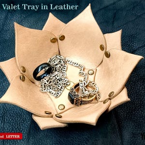 Op de afbeelding: Een beige leren lotusvormige valet tray met gouden klinknagels, met een zwarte ring, zilveren ketting en twee andere ringen. De tray heeft de tekst "Lotus Valet Tray in Leather" bovenaan. De achtergrond is een donkerblauwe, getextureerde ondergrond.