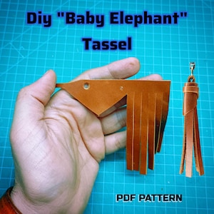 May include: A tan leather piece cut into a stylized elephant shape with a fringed tail and a small circular hole for an eye, alongside a finished tan leather tassel with a metal clasp. The text reads: Diy "Baby Elephant" Tassel PDF PATTERN.