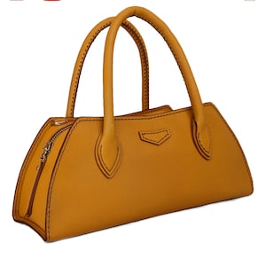 May include: A mustard yellow handbag with a semi-circular silhouette, two rounded top handles, and a side zipper closure. The bag features visible stitching along the edges and a small, shield-shaped decorative element on the front center.