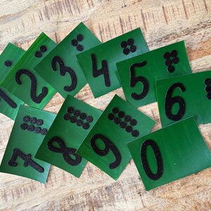 May include: Green number cards with black glitter numbers and corresponding dot patterns. The cards display numbers 0 through 9, ideal for educational activities. The cards are arranged on a wooden surface.