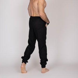 Könnte beinhalten: Schwarze Jogginghose mit elastischen Bündchen. Die Hose ist aus einem weichen Material. Das Model schaut über die Schulter. Der Hintergrund ist einfarbig hellgrau.