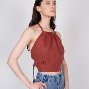 May include: A rust-colored halter top featuring a gathered neckline, thin straps that tie at the back, and a cropped hemline, paired with high-waisted blue denim jeans that have a gathered elastic waistband and a button-front closure.